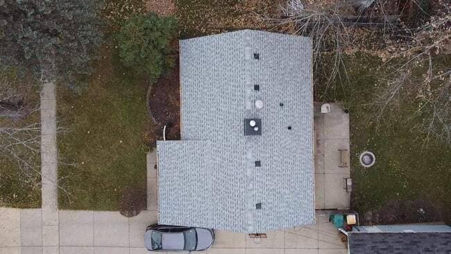 Direct overhead drone view of a completed light gray architectural asphalt shingle roof on a suburban home. The roof features a central chimney with clean flashing, four integrated roof vents, and a low-pitch design over a secondary structure. The image highlights the precise shingle placement and the professional finish of the roofing system.